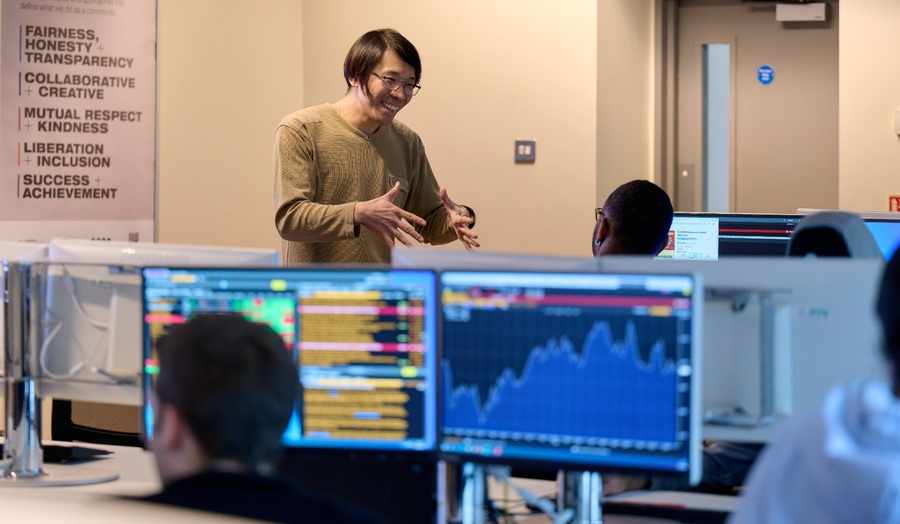 A person standing in a computer lab gestures while speaking to seated students at Bloomberg terminal workstations