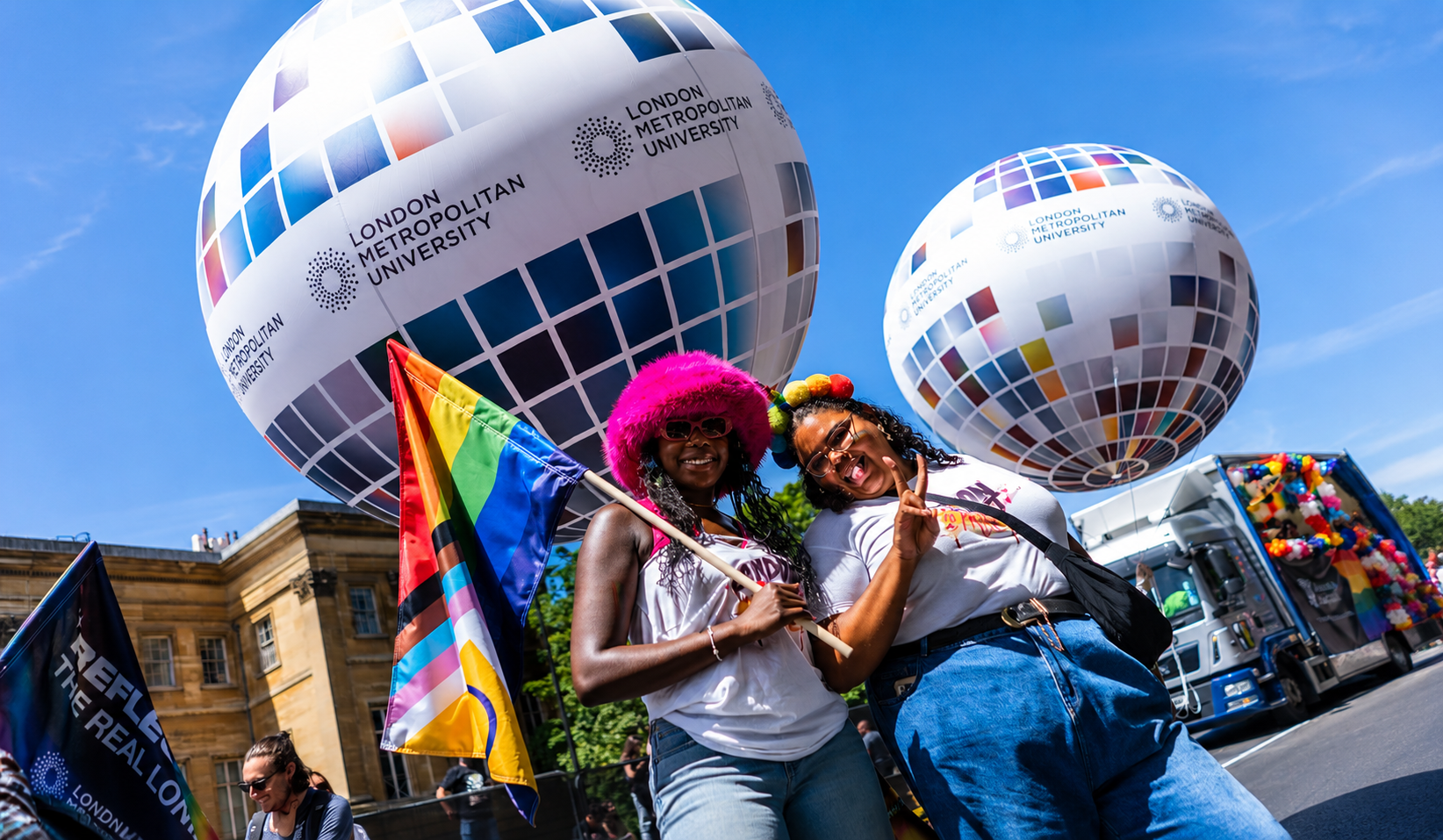 Two people smile and hold a Progress Pride flag beneath London Metropolitan University branded balloons at a Pride parade.