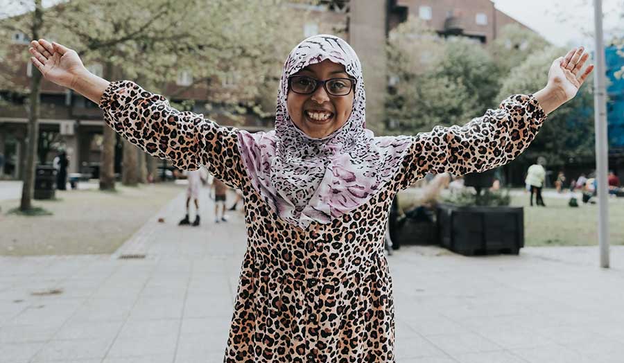 A woman, wearing a colourful headscarf and dress, smiles with her arms outstretched standing outside